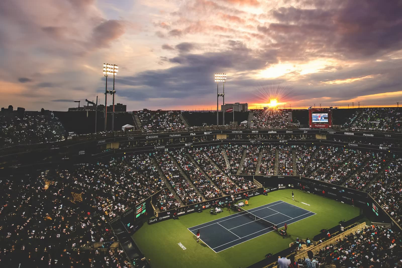 Tennis stadium at sunset with spectators and floodlit court.