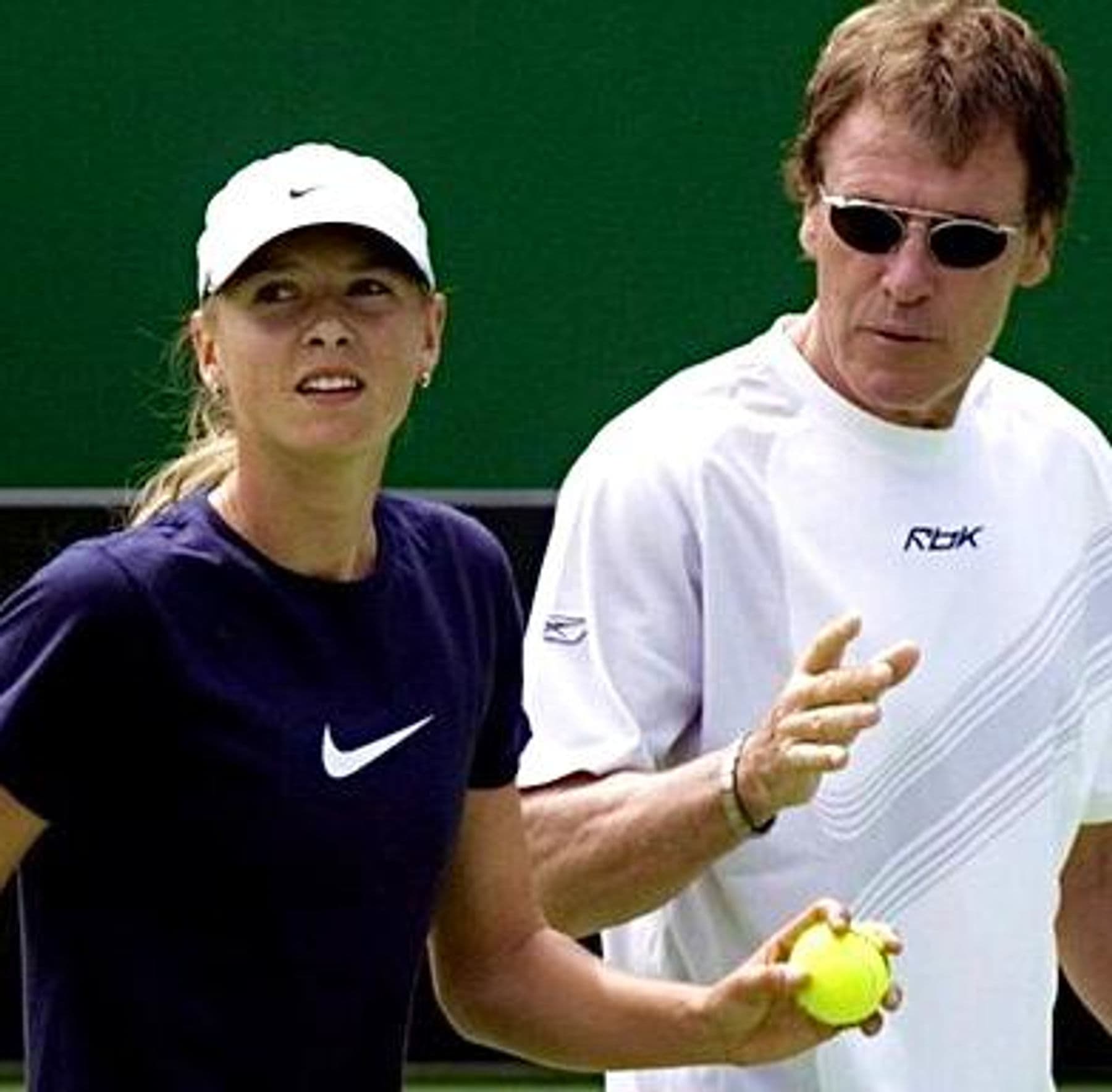 Tennis coach and player on an outdoor tennis court reviewing technique — green backdrop behind the baseline.