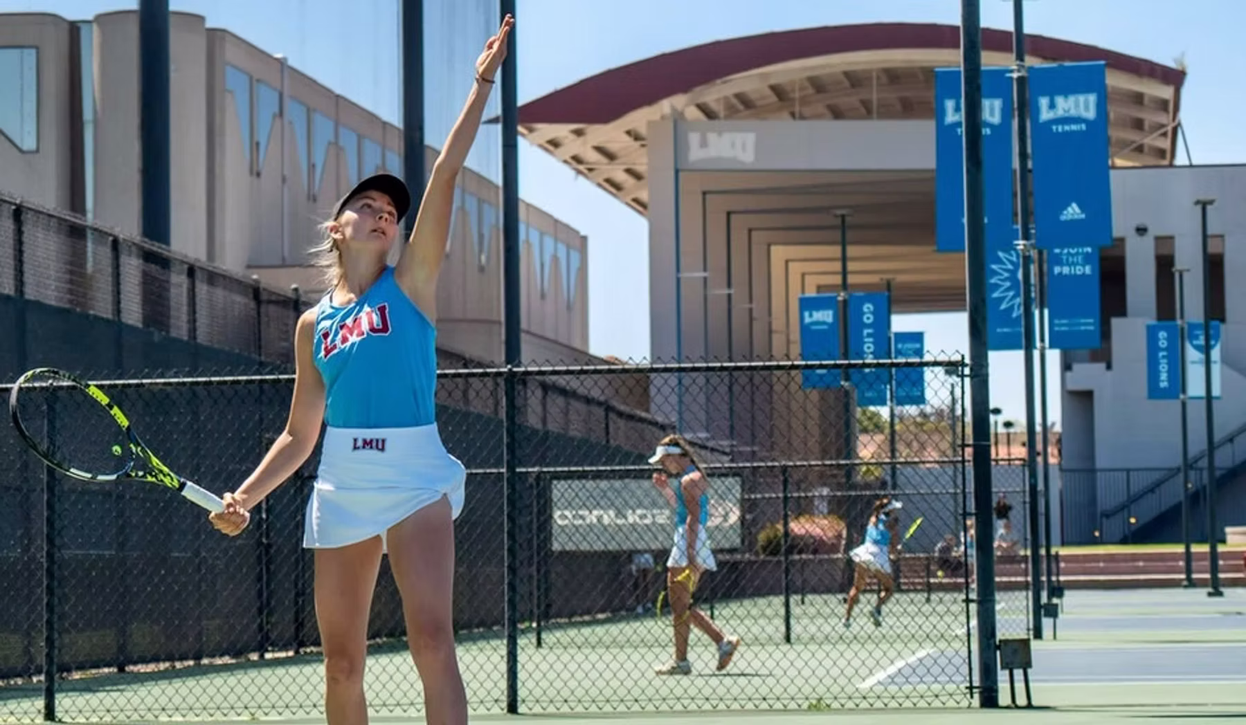 Tennis player in a college uniform serving on an outdoor court — campus banners visible behind the fence.