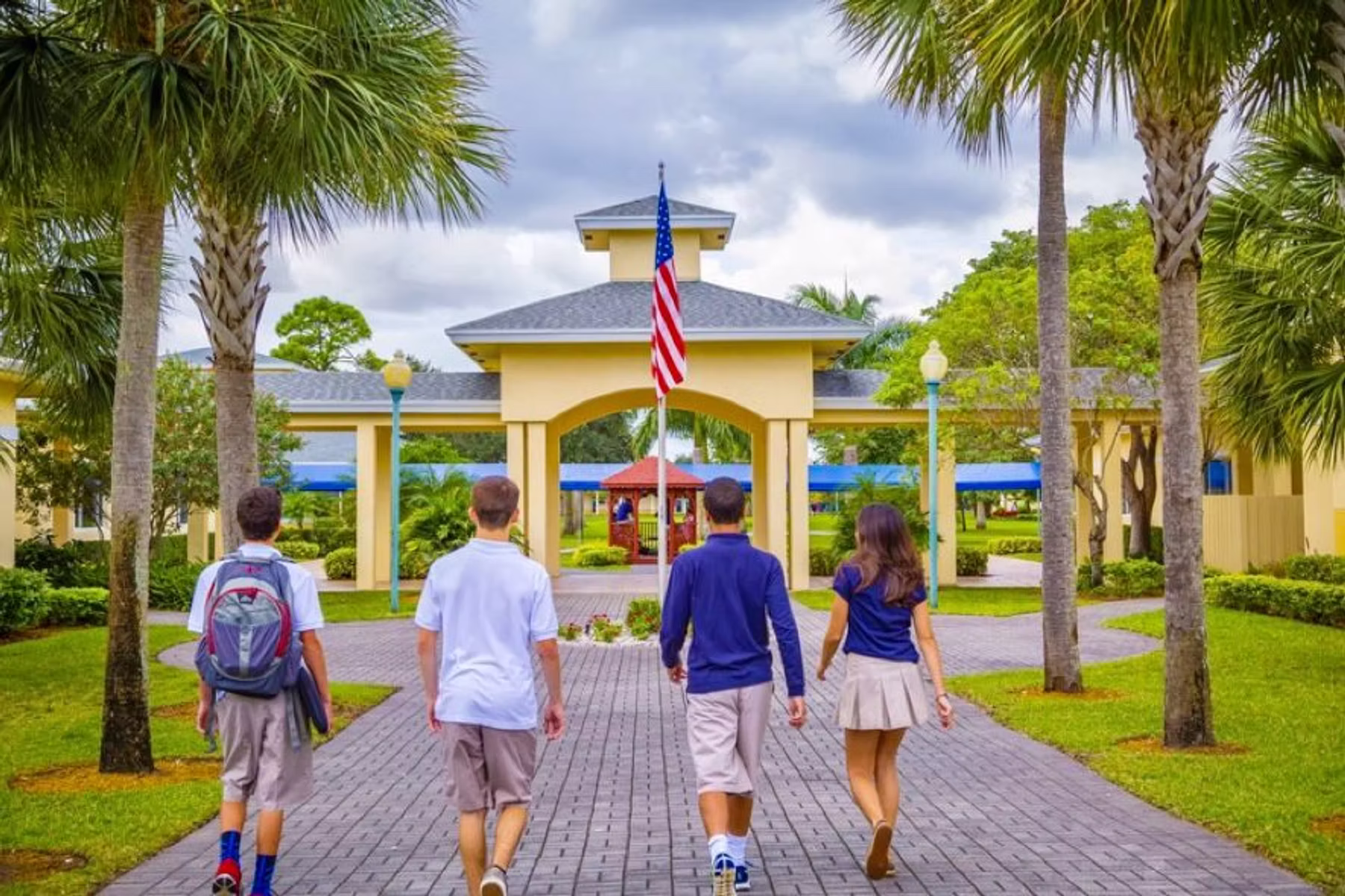 Four students walking on a paved path toward an American-flagged campus building with palms and lawns.